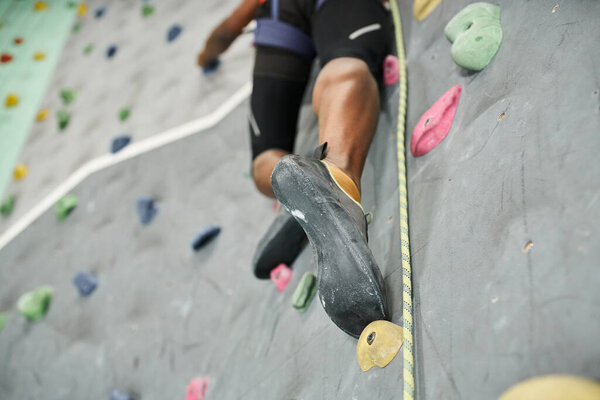 cropped view of legs of sporty african american man climbing up rock wall with safety rope