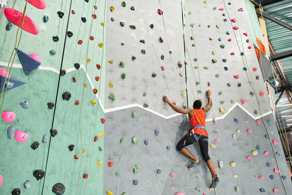 sporty african american man in orange shirt climbing up with alpine harness and safety rope