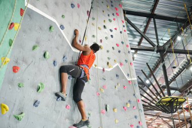 strong african american man in orange shirt using safety rope and alpine harness to climb up wall clipart
