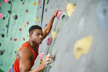 good looking african american man gripping on boulders using safety rope and looking at camera clipart