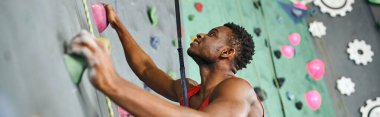 cheerful young african american man gripping on boulders while ascending up rock wall, banner clipart