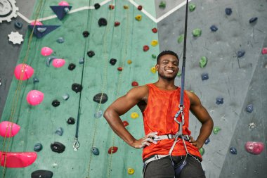 joyful young african american man posing with his arms akimbo and smiling cheerfully at camera clipart