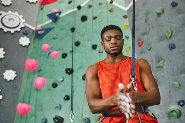 good looking african american man in orange shirt using talc powder before climbing up rock wall clipart