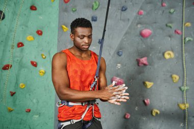 young athletic african american man in orange shirt with safety rope using talc powder, bouldering clipart