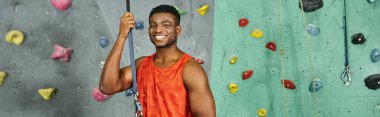 joyful athletic african american man in orange shirt smiling happily at camera, bouldering, banner clipart