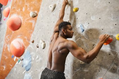 Atletik, Afro-Amerikalı, tişörtsüz, kaya duvarına tırmanan genç bir adamın arka görüntüsü.
