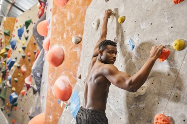 handsome shirtless african american man climbing up rock wall and looking at camera, bouldering clipart