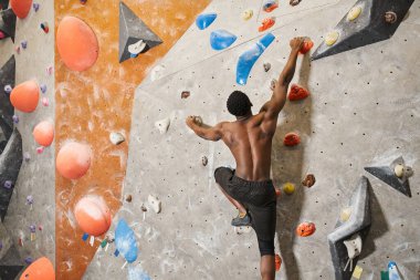 muscular shirtless african american man gripping strongly on boulders while climbing up rock wall clipart