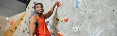 Turuncu tişörtlü neşeli Afro-Amerikalı adam kayalara tutunurken neşeli bir şekilde yüzünü çeviriyor.
