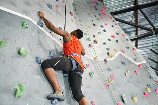 muscular african american man with alpine harness and safety rope climbing up bouldering wall