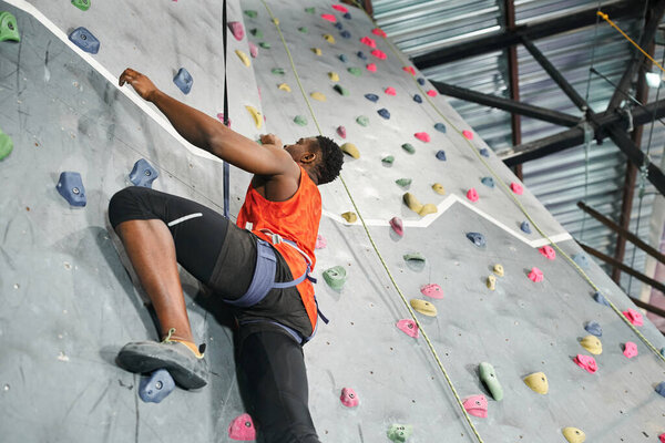 athletic young african american man in orange shirt using safety rope to climb up bouldering wall