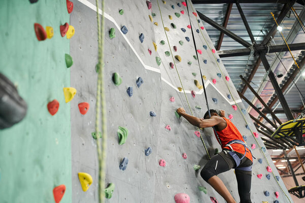 sporty young african american man in orange shirt ascending up bouldering wall with safety rope