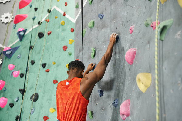 back view of african american man in orange shirt climbing up bouldering wall with safety rope