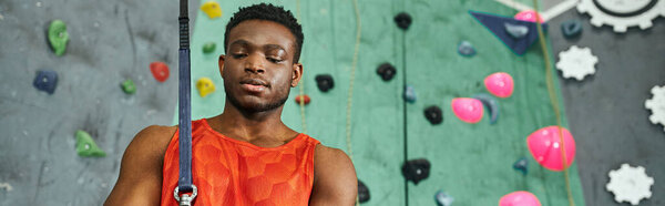 handsome sporty african american man posing with safety rope next to climbing wall, banner