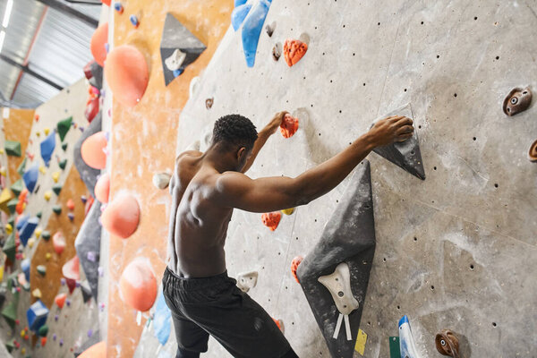 sporty topless african american man posing on bouldering wall and gripping on rocks, looking down