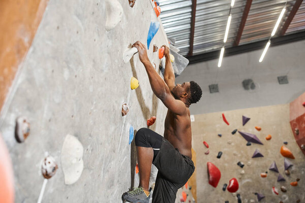 shirtless handsome african american man in black pants climbing up bouldering wall and looking up