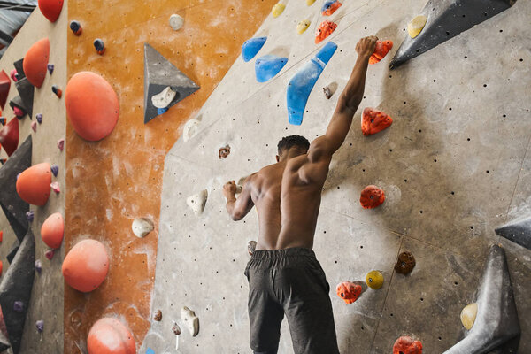 shirtless young african american man in black pants climbing up bouldering wall and looking down