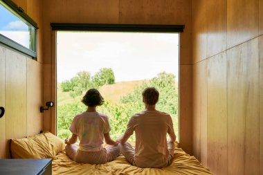 Birlikte meditasyon yapan çiftlerin arka planı ve orman manzaralı pencerenin yanındaki yatakta oturmaları.