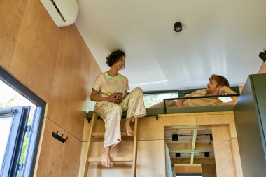 happy asian woman holding cup of coffee and sitting at cozy bunk bed with redhead boyfriend clipart