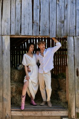 newlyweds in countryside, asian bride in cowboy boots and white dress standing with groom in barn clipart