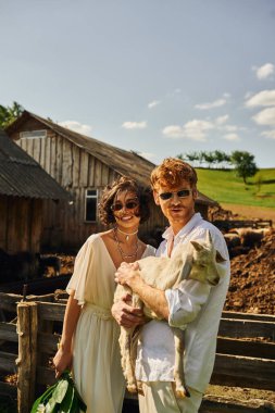 Boho tarzında kırsal bir düğün, neşeli damat Asyalı gelinlik içinde bebek keçiyi tutuyor.