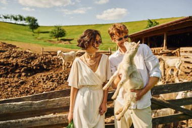 Yeni evli bir çift, kızıl saçlı damat beyaz elbiseli, boho tarzı bir Asyalı gelinin yanında bebek keçi tutuyor.