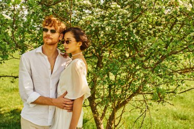 young multiethnic newlyweds in sunglasses and wedding gown hugging in green garden, boho style clipart