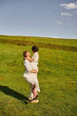 joyous newlyweds, happy groom lifting asian woman in white dress and cowboy boots in green field clipart
