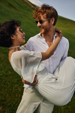 Mutlu yeni evliler, neşeli damat yeşil tarlada beyaz elbiseli Asyalı gelini kaldırıyor.