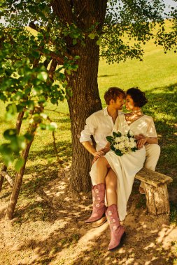 redhead groom embracing asian bride with bouquet while sitting on bench under tree, rustic wedding clipart