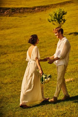 redhead groom and asian bride with flowers wearing elegant boho attire and holding hands in field clipart