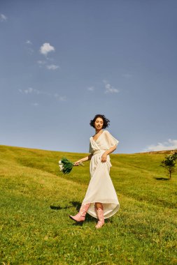 carefree asian woman in wedding dress and cowboy boots walking with flowers in green field