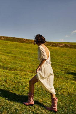 Beyaz elbiseli esmer kadın ve kovboy çizmeleri, mavi gökyüzünün altında yeşil tarlada yürüyor, kırsal alanda.