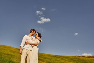 Boho tarzında ırklar arası yeni evliler. Mavi gökyüzünün altında yeşil alanda kucaklaşıyorlar. Kırsal bir düğün.
