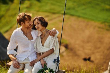 Mutlu kızıl damat yakışıklı Asyalı geline sarılıyor. Manzaralı kırsalda salıncakta salıncak sallıyor.