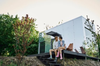 Modern çim evin verandasında oturan ve birbirlerine gülümseyen kaygısız ırklar arası bir çift.