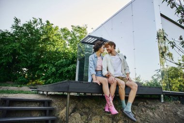 full length of happy interracial couple sitting on porch near glass house and looking at each other clipart