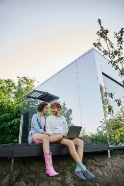 Bilgisayarlı mutlu ırklar arası çift kırsal kesimdeki modern cam evin verandasında oturuyor.