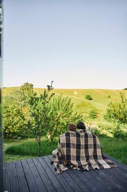 back view of young couple sitting on wooden porch under plaid blanket and enjoying scenic landscape clipart