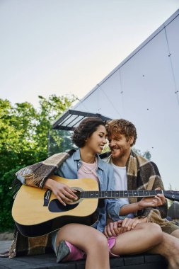 Mutlu Asyalı kadın kırsal kesimdeki cam evin yakınındaki verandada erkek arkadaşına akustik gitar çalıyor.
