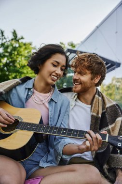 Neşeli Asyalı kadın kırsaldaki cam evin yakınındaki mutlu sevgiliye akustik gitar çalıyor.