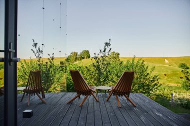 Modern cam evin yanındaki verandada manzaralı ahşap sandalyeler ve kahve masası.