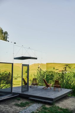 Çevre dostu cam ev, yeşil tepelerin yanında ahşap veranda manzaralı kırsal bölgede mavi gökyüzünün altında.