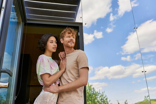 dreamy interracial couple standing at front door of modern glass house and looking away, fresh air