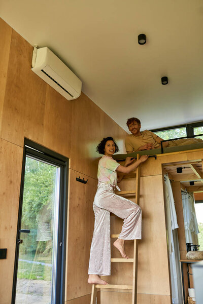 happy redhead man looking at camera while girlfriend climbing on ladder of bunk bed, weekend getaway