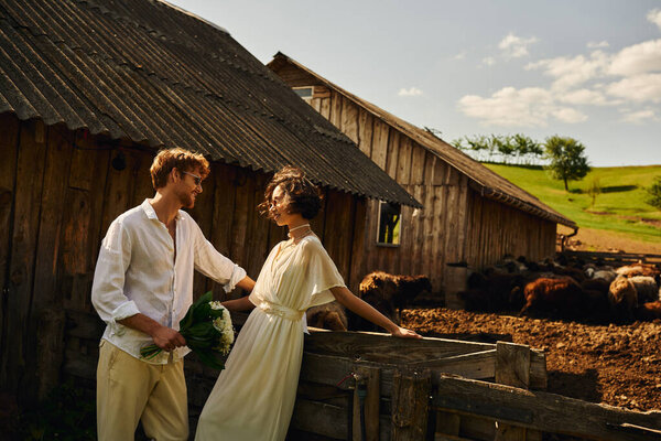 happy asian bride in wedding dress standing near groom in sunglasses near stable with sheep