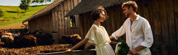 happy asian bride in wedding dress standing near groom in sunglasses near stable with sheep, banner