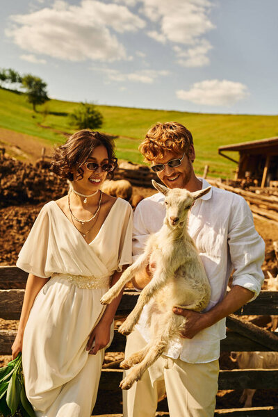 couple in countryside, happy groom holding baby goat near asian bride in white dress, boho style