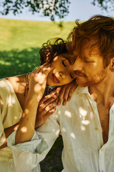 groom embracing asian bride in white dress and sitting under tree, sunlight and shadows on face