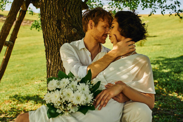 Рыжий жених обнимает элегантную азиатскую невесту, сидя с цветами под деревом, свадьба в сельской местности